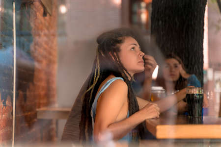 A young woman with dreadlocks sitting alone in a bar and drinking beer.