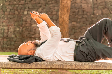 Asian businessman lying on a stone bench using mobile applications in a park in a metropolis.の写真素材