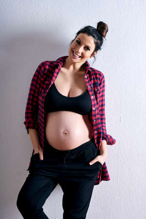 A young happy pregnant woman in a red shirt and a black underwear standing in front of a grey...