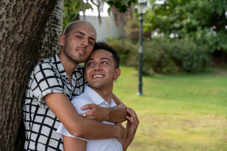 Happy Latin American gay couple embracing at a tree in a city park.の写真素材