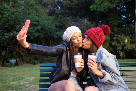 Friends taking a selfie and kissing with takeaway coffee sitting on a bench in a park.