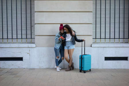Lesbian girls standing embracing and checking the phone on the street at the wall carrying a suitcase.の写真素材