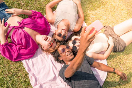 A group of multiethnic friends taking selfies while laying in the grass on a sunny day in a park.の写真素材