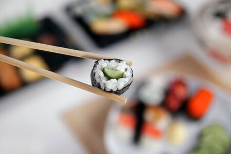 A pair of bamboo chopsticks holding a kappamaki with other sushi in the backgroundの写真素材