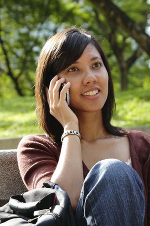 An Asian woman using a cellphone sitting on a bench at a public parkの写真素材