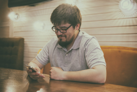 Young Beard Man in Glasses and Grey Shirt sitting in bar looking to the phone and smiling. Satisfied glad guy with smartphone in right hand.の写真素材