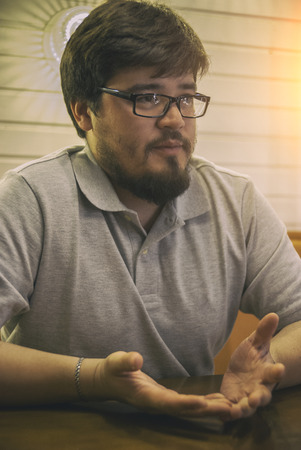 Young Beard Man in Glasses and Grey Shirt sitting in bar  and talking with partner. Glad guy