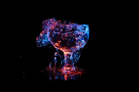 Glass Chalice overflowing with clear liquid under colored lights on a black backgroundの写真素材