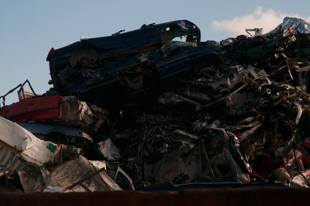 Photo of a heap of smashed cars and old metal in a junkyard with a blue sky and clouds in the...