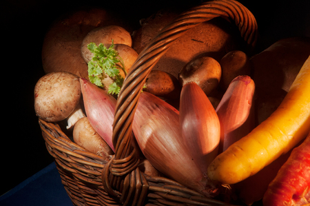 Wicker basket filled with mushrooms, onions and carrots with a sprig of green parsley in low key lighting on a black backgroundの写真素材