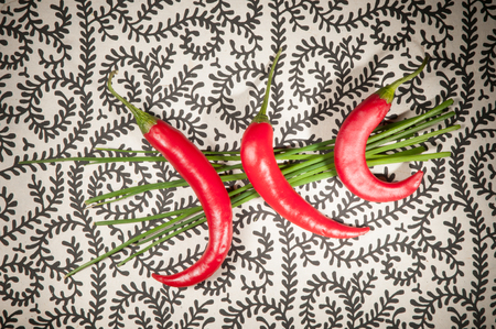Three bright red chili peppers on top of a small bunch of vibrant green chives all on a white background with  a swirly black patternの写真素材