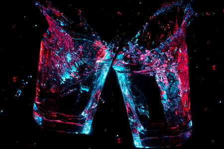 2 rocks glasses clinking a toast with colorful glowing liquid splashing from them on a black backgroundの写真素材