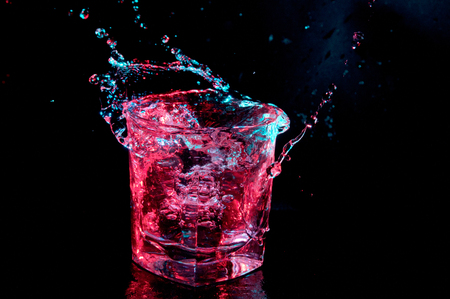 Water splashing out from a rocks glass in colored lights isolated on a black backgroundの写真素材