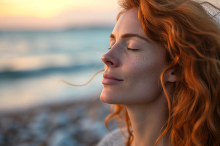 Serene auburn woman with closed eyes enjoying a seaside breeze at sunset, with soft focus background.の素材