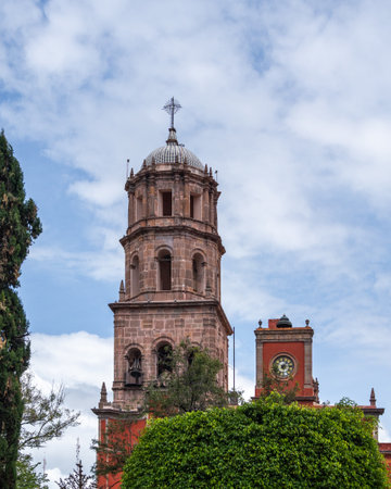 A tall tower with a cross on top and a clock on the side. The tower is surrounded by trees. Historic...