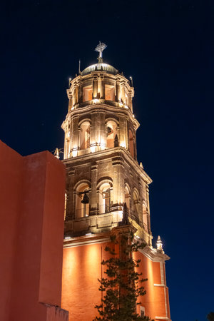 A tall tower with a cross on top is lit up at night. The tower is surrounded by a red building....