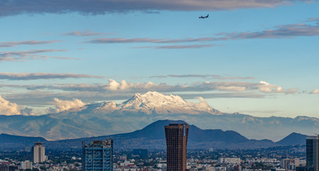 A mountain range is visible in the distance behind a city. A plane is flying over the city. Panoramic view of Mexico City, snow-covered volcanoes Popocatepetl and Iztaccihuatlの写真素材