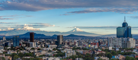 A city with a mountain range in the background. The city is full of tall buildings and the sky is blue. Panoramic view of Mexico City, snow-covered volcanoes Popocatepetl and Iztaccihuatlの写真素材