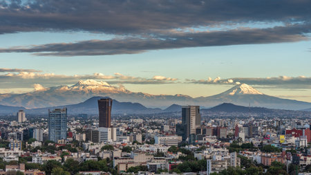 A city with a mountain range in the background. The sky is cloudy and the sun is setting. Panoramic view of Mexico City, snow-covered volcanoes Popocatepetl and Iztaccihuatlの写真素材