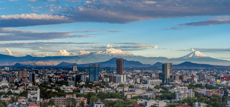 A city skyline with mountains in the background. The sky is blue with some clouds. Panoramic view of Mexico City, snow-covered volcanoes Popocatepetl and Iztaccihuatlの写真素材