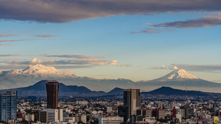 A city with mountains in the background. The sky is blue and the clouds are white. Panoramic view of Mexico City, snow-covered volcanoes Popocatepetl and Iztaccihuatlの写真素材