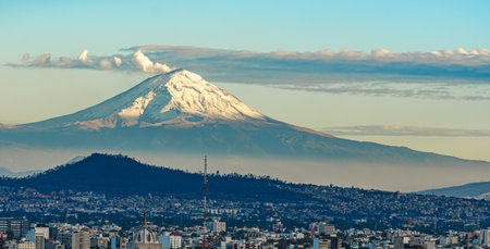A mountain with a snow covered peak is in the background of a city. The city is bustling with activity. Panoramic view of Mexico City, snow-covered volcanoes Popocatepetl and Iztaccihuatlの写真素材