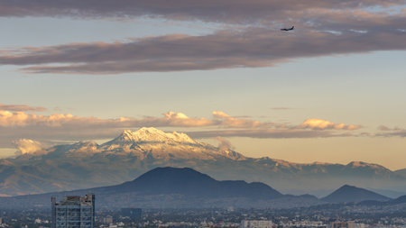 A mountain range is visible in the distance with a plane flying above it. The sky is cloudy and the sun is setting. Panoramic view of Mexico City, snow-covered volcanoes Popocatepetl and Iztaccihuatlの写真素材