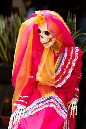 A skeleton dressed in a red dress with a veil and flowers. The skeleton is sitting on a chair. Mexican Day of the Dead tradition, offerings, food, colors, decorations, and papel picado, La Catrina.の写真素材