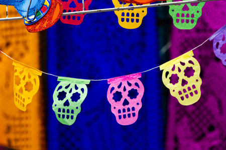 A colorful string of paper skulls hanging from a wire. The skulls are in various colors and sizes. Mexican Day of the Dead tradition, offerings, food, colors, decorations, and papel picado, La Catrina.の写真素材