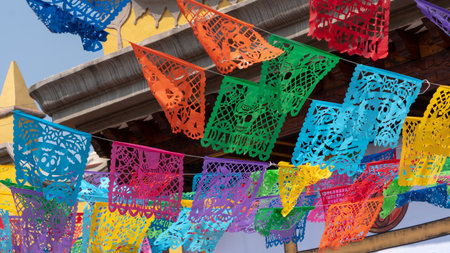 A colorful display of paper flags hanging from a building. The flags are of different colors. Mexican Day of the Dead tradition, offerings, food, colors, decorations, and papel picado, La Catrina.の写真素材