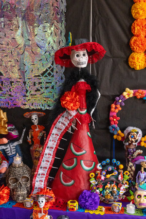 A woman dressed in a red dress and a black hat is standing in front of a table with many other figur. Mexican Day of the Dead tradition, offerings, food, colors, decorations, and papel picado.の写真素材