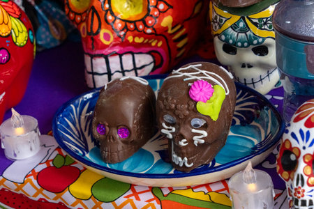 A plate of chocolate skulls with pink flowers on them sits on a table with other skulls and candles. Mexican Day of the Dead tradition, offerings, food, colors, decorations, and papel picado, La Catrina.の写真素材