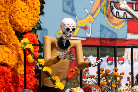 A skeleton is standing in front of a wall with a skull on it. The skeleton is holding a hand up. Mexican Day of the Dead tradition, offerings, food, colors, decorations, and papel picado, La Catrina.の写真素材