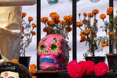 A pink skull is on a table next to some flowers. The skull is decorated with flowers and has a blue. Mexican Day of the Dead tradition, offerings, colors, decorations, and papel picado.の写真素材