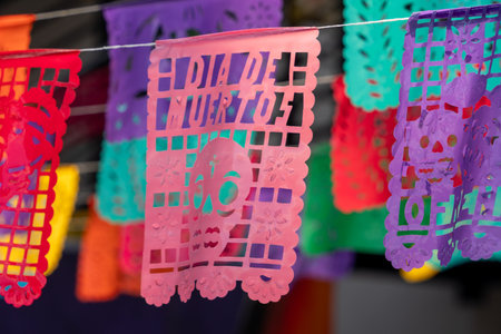 A bunch of colorful paper flags with skulls and the number 16 on them are hanging from a line. Mexican Day of the Dead tradition, offerings, food, colors, decorations, and papel picado, La Catrina.の写真素材