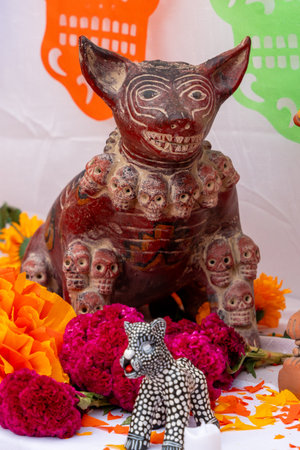 A small statue of a dog with a skull collar sits on a table with a bunch of flowers. Mexican Day of the Dead tradition, offerings, food, colors, decorations, and papel picado, La Catrina.の写真素材