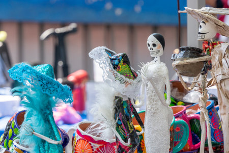 A table with many different types of skulls and skeletons. Some are dressed in clothing. Mexican Day of the Dead tradition, offerings, food, colors, decorations, and papel picado, La Catrina.の写真素材