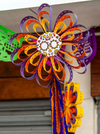 A colorful flower with a skull on it. The flower is made of paper and is hanging from a string. Mexican Day of the Dead tradition, offerings, food, colors, decorations, and papel picado, La Catrina.の写真素材