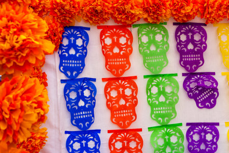 A vibrant display of paper skulls is arranged in a row, with orange flowers in the background. Mexican Day of the Dead tradition, offerings, food, colors, decorations, and papel picado, La Catrina.の写真素材