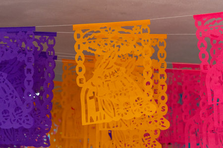 A colorful banner with a woman on it hangs on a string. The banner is purple, orange, and pink. Mexican Day of the Dead tradition, offerings, food, colors, decorations, and papel picado, La Catrina.の写真素材