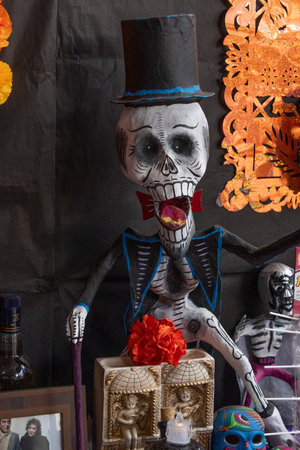 A skeleton dressed in a top hat and a cape is standing in front of a table with a vase and a bottle. Mexican Day of the Dead tradition, offerings, food, colors, decorations, and papel picado, La Catrina.の写真素材