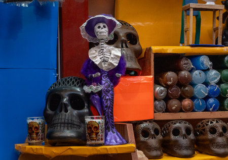 A table is filled with various skulls and skeletons, including a purple skeleton wearing a hat. Mexican Day of the Dead tradition, offerings, food, colors, decorations, and papel picado, La Catrina.の写真素材
