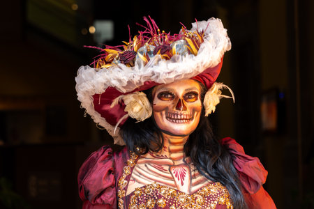 A woman is dressed in a red outfit with a skull painted on her face and a hat on her head. Mexican Day of the Dead tradition, offerings, food, colors, decorations, and papel picado, La Catrina.の写真素材