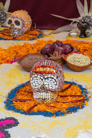 A skull made of beans is on a table with other items. The skull is surrounded by flowers. Mexican Day of the Dead tradition, offerings, food, colors, decorations, and papel picado, La Catrina.の写真素材