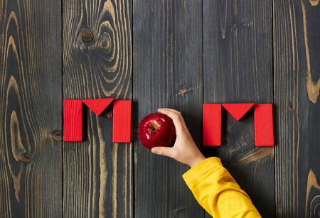 Mom word from red toy bricks and an apple with child hand flat on dark textured wooden background, copy space for your design, from above top view, Mother's day zero waste conceptの写真素材