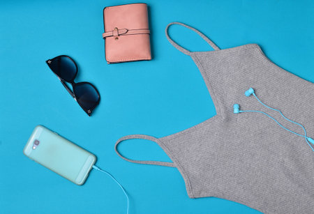 Women's clothing and accessories laid out on a blue background. Nightdress, T-shirt, purse, smartphone with headphones, sunglasses. Top View. Flat lay.の写真素材
