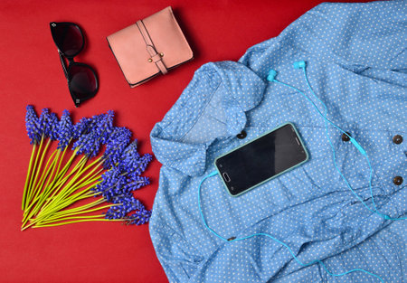 Women's clothing and accessories are lined on a red background.Shirt, smartphone, purse, sunglasses, headphones, blue wildflowers. Flat lay.の写真素材