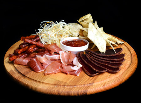 Snack to beer on a wooden board. Basturma, dried meat, dried squid, chips isolated on a black background.の写真素材