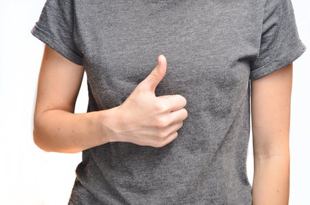 The thumb of a female hand raised upwards. A female trunk in a gray T-shirt is isolated on a white...