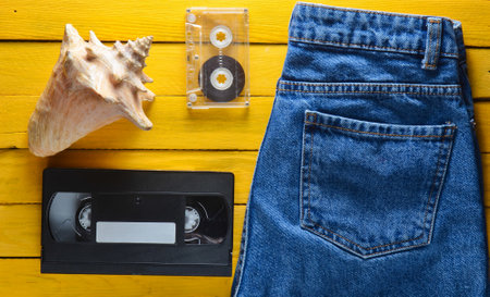 Accessories x generation: jeans, audio cassette, vhs, shell on a wooden table of yellow color....